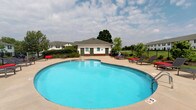Located at 2870 Kengary Way in Reynoldsburg, Ohio, this single-floor community amenity space is centered around a generously sized freeform outdoor swimming pool, making it an inviting hub for recreation and relaxation. The pool's curvilinear shape and clear blue water are complemented by a surrounding concrete deck outfitted with lounge chairs and accent seating, offering ample space for sunbathing and socializing.

A dedicated pool house anchors the space, featuring a classic exterior with white siding, dark green trim, and brick detailing. The structure provides essential support facilities for pool guests and is framed by lush, mature trees that lend a sense of privacy and natural beauty to the setting.

The entire pool area is enclosed by a black metal perimeter fence, ensuring both safety and a clean, defined boundary. Residential buildings visible in the background reflect a cohesive architectural style throughout the community, reinforcing the neighborhood's well-planned character.

The overall ambiance of this outdoor space is welcoming and functional, designed to serve as a central gathering point for residents. With open skies above and greenery surrounding the area, the space offers a refreshing retreat within the broader Reynoldsburg community.