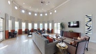 This single-floor lounge space makes an immediate impression with its dramatically high, curved ceilings accented by crown molding and a circular ring of porthole-style windows near the top, flooding the interior with natural light from multiple angles. A ceiling fan anchors the center of the room, while recessed lighting supplements the abundant daylight throughout. The walls are finished in a soft neutral tone, creating a clean, welcoming backdrop.

The main seating area is anchored by a large sectional sofa in light gray upholstery, accented with rust and coral throw pillows that echo the two-toned drapery panels framing the many tall windows. A dark wood media console along one wall supports a wall-mounted flat-screen television, while a tall potted tropical plant adds a natural, organic element nearby. Additional seating clusters near the windows offer more intimate conversation areas, furnished with gray wingback chairs and small accent tables.

Light wood-toned flooring runs throughout, contributing to the room's bright and airy atmosphere. Decorative accents, including a sculptural centerpiece on a gold side table, add character and visual interest, giving the space a polished, community-oriented ambiance.