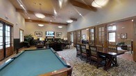 This single-floor community clubhouse offers a thoughtfully designed gathering space that seamlessly blends recreation, socializing, and relaxation. The centerpiece of the room is a full-size billiards table set atop a richly patterned carpet, lending the space a classic, upscale character. Vaulted ceilings with exposed wood beam accents soar overhead, amplifying the sense of openness and grandeur, while a combination of pendant lights and recessed fixtures casts a warm, inviting glow throughout.

A comfortable lounge area is arranged nearby, featuring plush seating, decorative table lamps, and framed artwork on the walls, creating an intimate nook perfect for conversation or unwinding. A wall-mounted flat-screen television adds a modern amenity to the traditional aesthetic. Adjacent to the lounge, a formal dining area with dark wood chairs and a long table accommodates group gatherings with ease.

French-style glass-paned doors line one wall, allowing natural light to filter in and offering views into adjoining spaces, while maintaining a sense of architectural elegance. The warm earth-toned walls, wood trim details, and layered lighting combine to create a refined yet comfortable ambiance suitable for a wide range of activities and social occasions.