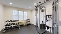 This fitness room offers a practical and well-organized workout space designed to accommodate a variety of exercise routines. The room features durable rubber flooring, which provides both safety and comfort during workouts. Natural light filters in through large windows fitted with horizontal blinds, creating a bright and welcoming atmosphere.

The space is thoughtfully outfitted with a comprehensive range of equipment. A multi-tiered dumbbell rack lines one wall, holding an extensive selection of free weights to support strength training at various levels. Adjacent to it, a second rack displays a collection of medicine balls, adding versatility for functional and core training exercises. A robust cable machine anchors the room, offering multiple attachment points and a weighted stack suitable for a wide range of resistance exercises.

Recessed ceiling lighting supplements the natural light, ensuring the space remains well-lit throughout the day. The white walls and clean layout contribute to an open, uncluttered feel. Overall, the fitness room is efficiently designed to provide residents with accessible, multi-functional workout options in a comfortable, well-maintained environment.