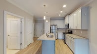 The Autumn is a well-appointed 1-bedroom, 1-bathroom unit designed with both function and style in mind. The kitchen serves as the heart of the home, showcasing white shaker-style cabinetry that extends to the ceiling, granite countertops, a classic subway tile backsplash, and a full suite of stainless steel appliances including a refrigerator, range, and microwave. A central kitchen island with a built-in sink and matching granite surface provides additional prep space and a natural gathering point, illuminated by sleek pendant lighting above. Light wood-look flooring runs throughout the main living and kitchen areas, complementing the bright, neutral color palette. The bedroom offers a comfortable retreat with plush carpeting, a ceiling fan with light fixture, and a window with blinds that invites natural light. The full bathroom features an extended granite-topped vanity with ample storage, a large mirror, and tile flooring, conveying a clean and functional design. A dedicated laundry area with wire shelving and washer/dryer hookups adds everyday convenience. Recessed lighting throughout enhances the overall brightness of the space, creating a welcoming and livable environment from entry to bedroom.