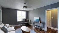 This single-floor space exudes a modern and refined aesthetic, anchored by a cohesive grey color palette that carries throughout the interior. The living area showcases rich, dark hardwood flooring that adds warmth and contrast against the cool-toned walls, creating a striking visual balance. A ceiling fan with a stylish industrial-inspired fixture serves as a functional centerpiece overhead, while natural light filters in through a window fitted with blinds, brightening the room and adding an airy feel. The space is thoughtfully furnished with a sleek oval coffee table, a contemporary grey armchair, and a metallic console table that doubles as a media stand. A landscape-inspired artwork leans against the console, introducing a subtle splash of color and artistic character to the room. A classic radiator beneath the window nods to the building's architectural heritage, blending seamlessly with the modern furnishings. The overall ambiance is clean, understated, and sophisticated, making this a comfortable yet stylish living environment well-suited for both relaxing and entertaining.