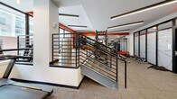 Located at 916 West St in Charlottesville, Virginia, this single-level fitness center offers a well-appointed workout environment designed with both functionality and modern aesthetics in mind. The space is defined by clean lines, a neutral color palette punctuated by bold orange accent walls, and durable rubber-composite flooring suited for high-traffic athletic use.

The main workout area is generously sized, accommodating multiple Matrix elliptical trainers and cardio machines arranged across an open floor plan. A dedicated functional training zone features a multi-station cable and weight rack system, a heavy bag, and additional strength equipment positioned against the vibrant orange accent wall, creating a visually energetic training environment.

Frosted glass panel partitions with a geometric line design divide sections of the space, adding a refined architectural detail while maintaining an open, airy feel. Linear LED lighting runs across the ceiling, ensuring consistent, bright illumination throughout. Built-in blue shelving units and a water station are conveniently integrated along one wall. The entrance features frosted glass doors with matching geometric patterns. A stairwell with sleek black metal railings connects to adjacent areas of the building, contributing to the facility's polished, contemporary character.