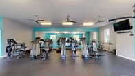 Located at 1315 Morreene Rd in Durham, North Carolina, this well-equipped fitness center offers a generously sized, single-floor exercise space designed to support a comprehensive workout experience. The room is finished with durable rubber flooring throughout, providing both safety and comfort underfoot during exercise.

The space is defined by its bold teal accent walls, which lend a vibrant, energizing atmosphere, complemented by crisp white ceilings and walls. Multiple ceiling fans are distributed across the facility, ensuring consistent air circulation and comfort during workouts. Natural light pours in through several windows, including a distinctive arched window, offering views of the surrounding greenery and keeping the environment bright and welcoming.

The fitness center is comprehensively outfitted with a wide variety of equipment. A dedicated cardio zone features treadmills and stationary bikes, while a strength training area includes multiple selectorized cable and resistance machines, a multi-functional training tower, and a free weights section stocked with dumbbells and benches. A wall-mounted television is available for entertainment, and a water fountain offers convenient hydration. Large mirrors along one wall enhance the sense of space and allow users to monitor their form. The layout is thoughtfully organized into distinct zones, making the facility functional and easy to navigate.