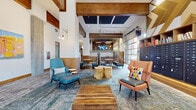 This single-floor communal space showcases a bold, contemporary design that blends industrial and mid-century modern aesthetics. Soaring ceilings with exposed wood beams define the expansive main lounge, which features a patterned area rug anchoring an eclectic mix of seating — including teal lounge chairs with ottomans, an orange accent chair with colorful throw pillows, and natural wood stump side tables alongside a slatted wood coffee table. Concrete-look columns and chevron-patterned wood wainscoting add architectural interest throughout.

A dining and gathering area features semi-private booth seating upholstered in vibrant floral fabric, framed by a dramatic geometric accent wall in gold and white. Overhead, acoustic ceiling panels and a chandelier lend both function and elegance to the space.

The communal kitchen is well-appointed with warm wood cabinetry, brass hardware, a large dark stone-topped island, stainless steel appliances, and white tile backsplash beneath a bold mural. Pendant lighting in gold tones adds warmth. A dedicated mail and package area with built-in lockers and shelving is conveniently integrated near the lounge. Colorful original artwork displayed throughout contributes to the space's vibrant, curated atmosphere.