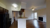 This single-story unit offers a practical and comfortable layout designed to maximize its well-proportioned square footage. The open-concept design seamlessly connects the kitchen and living areas, creating a cohesive and functional living space.

The kitchen features dark wood cabinetry paired with granite-look countertops and a peninsula that doubles as a breakfast bar, providing additional seating and workspace. A decorative upper cabinet structure adds architectural interest and helps define the kitchen zone within the open floor plan.

The living area is finished with rich, dark hardwood-style flooring that extends throughout the main living spaces, adding warmth and visual continuity. Crown molding frames the ceiling, while a ceiling fan with integrated lighting provides both comfort and illumination. A fireplace serves as a focal point along one wall, adding a cozy ambiance to the room. A window with blinds allows natural light to filter into the space.

The bathroom is accessible from the main living area and features a toilet and basic fixtures visible from the hallway. Overall, the unit presents a clean, move-in-ready environment with classic finishes throughout.