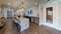 This single-floor amenity space blends sophisticated design with everyday functionality, making it an inviting destination for work, relaxation, and socializing. The open layout is unified by herringbone-patterned flooring that transitions seamlessly between areas, complemented by crisp crown molding and recessed lighting throughout.

The lounge area serves as the centerpiece of the space, furnished with elegantly patterned armchairs accented by chartreuse throw pillows, arranged around a sleek round coffee table. Bold abstract artwork in black, white, and green tones adorns the walls, adding a vibrant artistic energy to the room. Floor-to-ceiling glass partitions and large street-facing windows flood the space with natural light while offering views of the surrounding area, further enhanced by cascading pendant chandeliers that provide warm, ambient illumination.

Adjacent to the lounge, a dedicated business workstation features a long, dark wood desk fitted with dual monitors and office seating, set against a geometric textured accent wall — ideal for productivity in a polished environment.

The wet bar area is a standout feature, anchored by a generously sized granite-topped island with bar stool seating. Pendant lighting hangs above the island, while the back counter hosts a wall-mounted television, a built-in beverage refrigerator, a sink, and ample cabinetry — all finished in rich wood tones against a white tile backsplash.