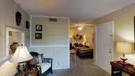 This single-story space showcases a thoughtful layout connecting a sitting area and bedroom. A prominent white shiplap half-wall serves as a defining architectural feature, creating a visual boundary between the two areas while maintaining an open feel. The sitting area accommodates a wicker chair with cushioned seating, complemented by soft lighting from a nearby lamp. Decorative wall art adorns the shiplap paneling, adding character to the space. Beyond the dividing wall, the bedroom features plush carpeted flooring that extends throughout, providing warmth and comfort underfoot. A ceiling fan with an integrated light fixture anchors the bedroom ceiling, offering both ventilation and ambient illumination. The bedroom is furnished with a full-sized bed dressed in layered, earth-toned bedding, flanked by a bedside lamp that casts a warm glow. Decorative wall accents above the bed add a personalized touch. A closet with sliding doors is conveniently positioned near the bedroom entrance, providing practical storage. The warm, neutral wall tones throughout create a cohesive and inviting atmosphere across both areas of this space.
