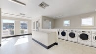 This single-floor shared laundry facility is designed for efficiency and ease of use, offering a spacious, open layout with all the essentials for a complete laundry experience. The room is outfitted with a generous row of front-loading washing machines along one wall, providing ample capacity for high-volume use. Along the adjacent wall, multiple stacked dryer units maximize the available space while keeping the floor area open and easy to navigate. A built-in folding counter runs along the perimeter, offering a convenient surface for sorting and folding laundry. The space is finished with durable light-toned vinyl tile flooring and neutral-painted walls, creating a clean and utilitarian aesthetic. Overhead fluorescent lighting ensures the room is well-lit throughout, while several windows allow natural light to fill the space and provide views of the surrounding outdoor area. A central dividing counter island adds additional workspace and helps organize the flow of the room. The facility also includes a dedicated electrical panel and thermostat, reflecting thoughtful infrastructure planning. Overall, the space is functional, bright, and well-maintained, making it a practical and accessible amenity.
