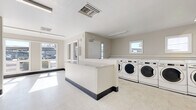 This single-floor shared laundry facility is designed for efficiency and ease of use, offering a spacious, open layout with all the essentials for a complete laundry experience. The room is outfitted with a generous row of front-loading washing machines along one wall, providing ample capacity for high-volume use. Along the adjacent wall, multiple stacked dryer units maximize the available space while keeping the floor area open and easy to navigate. A built-in folding counter runs along the perimeter, offering a convenient surface for sorting and folding laundry. The space is finished with durable light-toned vinyl tile flooring and neutral-painted walls, creating a clean and utilitarian aesthetic. Overhead fluorescent lighting ensures the room is well-lit throughout, while several windows allow natural light to fill the space and provide views of the surrounding outdoor area. A central dividing counter island adds additional workspace and helps organize the flow of the room. The facility also includes a dedicated electrical panel and thermostat, reflecting thoughtful infrastructure planning. Overall, the space is functional, bright, and well-maintained, making it a practical and accessible amenity.