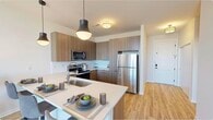 This single-floor residence showcases a thoughtfully designed open-concept layout with a contemporary aesthetic throughout. The kitchen serves as a central hub, featuring warm wood-grain cabinetry that extends to the ceiling for ample storage, stainless steel appliances, and a generous peninsula with a white quartz countertop that accommodates bar seating. Pendant lighting above the peninsula adds a stylish focal point, while light wood-style flooring ties the kitchen seamlessly into the adjoining living area. The living space is bright and airy, benefiting from expansive floor-to-ceiling windows that flood the room with natural light and offer open outdoor views. The bedroom continues the clean, modern feel with neutral tones, carpeted flooring, a ceiling fan, and its own access to natural light through large windows. A spacious walk-in closet with built-in shelving and hanging rods provides generous storage. The bathroom is finished with a wood-grain vanity, white quartz countertop, large mirror, and a glass-enclosed walk-in shower, all set against light tile flooring. Throughout the unit, a consistent palette of whites, warm wood tones, and soft neutrals creates a cohesive and inviting atmosphere.