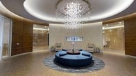Located at 201 Willett Ave in Port Chester, New York, this sophisticated lobby space makes a striking first impression with its thoughtfully curated contemporary design. The centerpiece of the room is a breathtaking cascading chandelier composed of spherical glass bubbles, suspended from a recessed circular ceiling detail that adds architectural depth and draws the eye upward. Warm wood-toned flooring and neutral stone-clad walls create an inviting, upscale atmosphere throughout the space. At the heart of the lobby sits a circular sectional sofa in deep navy blue, arranged around a round dark wood coffee table and anchored by an elegant area rug with intricate patterning — ideal for welcoming guests or encouraging conversation. A sleek linear gas fireplace is recessed into the feature wall, adding warmth and a refined focal point. Flanking the seating area are two accent chairs with side tables, completing the balanced, symmetrical layout. A decorative glass or backlit panel element is visible along one side wall, adding a luminous, artistic touch. The overall ambiance is polished and modern, combining luxurious materials with functional design to create a welcoming and impressive entrance experience.