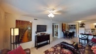 This single-floor residence offers a thoughtfully designed open-concept layout that seamlessly connects the living and dining areas, creating a cohesive and functional living environment. The living room serves as the centerpiece of the home, featuring light-toned flooring, a textured ceiling, and a ceiling fan with integrated lighting that provides both comfort and illumination. A wall-mounted flat-screen television is positioned above a dark wood console, adding a modern touch to the space. Warm, earthy tones are carried throughout via decorative artwork and an area rug with rich botanical patterns, contributing to an inviting and sophisticated atmosphere. The dining area, visible from the living room, accommodates a full dining set with upholstered seating, making it well-suited for entertaining. A glimpse into the hallway reveals access to additional rooms, suggesting a well-organized floor plan. Natural light filters through the space, enhancing the overall brightness and openness of the interior. The consistent use of warm accent colors and coordinated furnishings gives the residence a cohesive, well-curated aesthetic throughout.