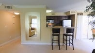This single-floor space is thoughtfully designed to maximize functionality and visual openness. The kitchen is a standout feature, outfitted with dark espresso-toned cabinetry that extends to the upper walls, providing ample storage. A stainless steel refrigerator and built-in microwave complement the cabinetry, while light-toned countertops offer a subtle contrast to the darker wood tones. A raised breakfast bar extends from the counter, accompanied by two dark-framed bar stools with cushioned seats, creating a casual dining or workspace area that also serves as a natural divider between the kitchen and living areas.

The living area is finished with neutral wall-to-wall carpeting and warm beige walls, contributing to a calm and cohesive atmosphere throughout. A large floor-length mirror with a white frame is mounted along one wall, effectively reflecting light and creating a greater sense of depth and openness. Crown molding runs along the ceiling, adding a polished architectural detail. The overall design balances practicality with a clean, comfortable aesthetic suited to everyday living.