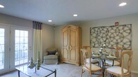 This single-floor space presents an open-concept layout that seamlessly integrates a living area and dining space. The room is finished with neutral-toned walls and plush carpeting, creating a warm and inviting atmosphere throughout. Recessed ceiling lights provide consistent, ambient illumination across the space, complementing the textured ceiling above.

The living area features a cushioned armchair with a decorative pillow, positioned beside a large, ornately carved wooden armoire with detailed paneling — a standout furniture piece that adds a classic, traditional character to the room. Patterned curtains in muted green and grey tones frame a window, softening the space with natural light.

Transitioning into the dining area, a glass-top table is surrounded by elegantly carved wooden chairs with upholstered seats. A decorative metal wall art piece with an interlocking circular design anchors the dining space visually, while a centerpiece arrangement on the table adds a refined finishing touch. Together, the furnishings and finishes reflect a cohesive, classically inspired interior design aesthetic.