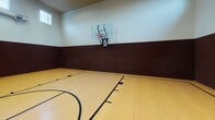 Located at 5701 Saratoga Blvd in Corpus Christi, Texas, this single-level indoor basketball facility offers a dedicated sports environment designed for athletic performance and recreation. The centerpiece of the space is a full hardwood basketball court, finished in a warm natural maple tone with crisp black court markings that include a center circle, key areas, and boundary lines — all consistent with regulation standards. A wall-mounted glass backboard with a standard hoop is positioned at one end of the court, providing a professional-grade playing experience. The walls feature a two-tone design, with a deep brown padded wainscoting lining the lower half to enhance player safety, transitioning to a light cream-colored upper wall. The facility boasts notably high ceilings, contributing to an open and airy atmosphere well-suited for active play. A row of clerestory windows positioned near the ceiling allows natural light to filter in, supplemented by overhead fluorescent lighting for consistent illumination. Visible HVAC ductwork and ventilation units are mounted along the upper walls, ensuring proper climate control throughout the space. Two exit doors are located on one end wall, providing easy access in and out of the court area. Overall, the facility presents a clean, functional, and well-maintained environment ideal for basketball and other court-based activities.