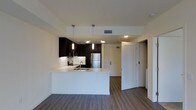 This single-floor residence at 100 Citronica in Lemon Grove showcases a clean, modern aesthetic with thoughtful design details throughout. The open-concept kitchen serves as a central highlight, featuring dark espresso cabinetry that creates a striking contrast against bright white countertops and walls. A peninsula with a built-in sink extends into the living area, offering both additional workspace and a casual seating option, making it ideal for entertaining or everyday use. Stainless steel appliances, including a refrigerator and range, lend a polished, contemporary feel to the space. Pendant lighting hangs above the peninsula, providing focused illumination while adding a stylish design touch. The flooring throughout is a warm-toned wood-look material that adds texture and continuity across the open layout. Smooth white walls and a clean ceiling with recessed vents contribute to the overall bright, airy atmosphere. A louvered door near the entry adds a classic architectural detail, while the open sightlines from the kitchen to the surrounding areas enhance the sense of spaciousness and functional flow within this well-appointed residence.