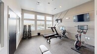 This fitness center in Box Elder, South Dakota, is a well-appointed, single-level exercise space designed to maximize functionality within a compact footprint. The interior is finished in a neutral gray palette, creating a clean, modern aesthetic throughout. A standout architectural feature is the geometric panel accent wall, which adds visual interest and serves as the backdrop for two wall-mounted flat-screen televisions, supporting both entertainment and workout tracking during exercise sessions.

The space is equipped with a variety of cardio and strength training equipment, including a treadmill, rowing machine, stationary bike, adjustable weight bench, a rack of dumbbells, and a medicine ball storage tower. Rubber-textured flooring provides durability and safety underfoot. Recessed ceiling lighting, combined with large windows along one wall, ensures the space is bright and well-illuminated. The windows offer direct views of an outdoor pool area, enhancing the overall ambiance with natural light and outdoor scenery.

A wall-mounted mirror adds depth to the space, while an interior pass-through window connects the fitness center to an adjacent common area. A dark-finished door provides a dedicated entry point, contributing to a polished, cohesive design throughout the facility.