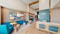 Located at 23350 North 23rd Avenue in Phoenix, AZ, this single-floor community lounge and lobby space at Bloom at Deer Valley is a thoughtfully designed gathering area that blends modern aesthetics with welcoming functionality. Soaring double-height ceilings amplify the sense of openness throughout, while wide-plank light hardwood flooring provides a warm, cohesive foundation underfoot.

The reception area greets visitors with a sleek white desk positioned against a bold teal accent wall bearing the Bloom at Deer Valley logo—immediately establishing the space's vibrant color palette. Adjacent to the reception, a fully equipped kitchenette bar features warm wood cabinetry, a patterned green tile backsplash, and a central island with seating for several guests, all illuminated by contemporary globe pendant lights.

Multiple seating zones define the expansive lounge, including a teal sofa grouping anchored by a large-scale abstract artwork and a curved blue sectional oriented toward a built-in media wall with open shelving adorned with greenery. A dedicated conference or dining table with mixed seating offers a versatile workspace. Floor-to-ceiling glass doors flood the interior with natural light and provide direct views of and access to the outdoor amenity area. Gold architectural beams add a refined finishing touch to this polished, community-centered space.