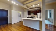 This well-appointed urban residence blends industrial-chic design with modern finishes across a single-floor layout. The open-concept kitchen serves as the heart of the home, showcasing rich dark cabinetry, a full suite of stainless steel appliances including a refrigerator, electric range, and built-in microwave, all centered around a generous white-topped peninsula with an integrated sink. Exposed ductwork runs overhead, adding an industrial character, while track lighting illuminates the warm hardwood floors that extend throughout the living area. A dedicated in-unit laundry area with a stacked washer and dryer provides everyday convenience just steps from the kitchen. The bedrooms are carpeted for comfort and feature large black-framed windows that frame views of the surrounding urban architecture, filling the rooms with natural light. One bedroom includes a walk-in closet with built-in rod storage. Two full bathrooms are finished consistently with dark wood vanities, white countertops, light tile flooring, and chrome fixtures; one bathroom offers a tub-shower combination with classic white subway tile surround, while the other provides a more compact, functional layout. The overall design balances urban industrial character with clean, contemporary residential finishes throughout.