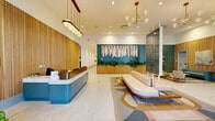 This thoughtfully designed lobby in New Orleans, LA offers a striking first impression through a cohesive blend of contemporary and mid-century modern aesthetics. The spacious single-floor layout is anchored by polished terrazzo flooring that reflects natural light streaming through large glass entry doors, creating a bright and airy atmosphere throughout.

A prominent reception desk finished in a muted teal blue with a clean white countertop serves as the functional centerpiece near the entrance, set against a backdrop of warm, vertically slatted wood wall paneling. Opposite the entry, a teal accent wall displays a striking macramé art installation above a built-in wood planter filled with lush greenery, adding texture and organic warmth.

Tall columns clad in deep blue-green elongated ceramic tile rise toward the high ceilings, creating dramatic vertical focal points that divide the space into distinct zones. A comfortable seating area features a low-profile cream sofa positioned atop a bold geometric area rug in muted earth tones. A secondary lounge nook with teal upholstered chairs and an arched display niche provides additional gathering space.

Overhead, a mix of globe-and-brass sputnik chandeliers and cylindrical pendant fixtures provide layered, ambient illumination. Potted plants are thoughtfully distributed throughout, enhancing the welcoming and vibrant ambiance of the space.