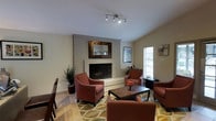 This single-story space centers around a well-appointed lounge area designed for comfort and casual gathering. The focal point of the room is a full brick fireplace, painted in a light neutral tone and topped with a decorative framed mirror that adds depth and visual interest to the wall. Track lighting mounted on the ceiling provides adjustable, direct illumination throughout the space.

The seating arrangement consists of several rust-toned upholstered armchairs positioned around a dark wood coffee table, creating an inviting conversational grouping. A bold, large-scale botanical-patterned area rug in earthy greens and yellows anchors the seating area and adds a lively contrast to the neutral walls and light tile flooring.

Natural light enters through a window fitted with horizontal blinds, brightening the space and complementing the warm interior tones. A tall potted plant in the corner adds a touch of greenery. The overall ambiance is warm, relaxed, and functional, making this space well-suited for lounging or informal meetings.