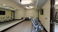 This single-level fitness room is thoughtfully designed to support a focused workout experience. The space is outfitted with a row of stationary cycling bikes arranged along one side, providing ample room for group or individual cycling sessions. A rack of kettlebells and free weights is positioned against the back wall, offering options for strength and conditioning exercises.

A large floor-to-ceiling mirror spans one wall, enhancing the sense of space while allowing users to monitor their form during workouts. The light-toned laminate flooring adds a clean, modern aesthetic and is practical for high-traffic fitness use. Dark wood baseboards provide a subtle contrast to the neutral, cream-colored walls.

Overhead, two ceiling fans with integrated lighting fixtures ensure proper air circulation and ventilation throughout the room. A wall-mounted flat-screen television is positioned prominently, ideal for streaming workout content or entertainment during training sessions. Recessed ventilation contributes to a comfortable indoor climate. Overall, the space maintains a clean, functional, and welcoming atmosphere suited for daily fitness routines.