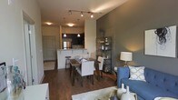 This single-floor residence offers a thoughtfully designed open-concept layout that brings together the living, dining, and kitchen areas in one cohesive, flowing space. Rich hardwood floors run throughout, anchoring the contemporary aesthetic and adding warmth to the interior.

The kitchen is appointed with dark cabinetry, stainless steel appliances, and a light-toned breakfast bar with counter seating — ideal for casual dining or entertaining. Pendant lighting above the bar adds a refined touch, while additional track lighting illuminates the main living and dining areas with versatility and style.

The dining area comfortably accommodates a full table and seating for several guests, positioned to take advantage of the open sightlines across the space. Just beyond, the living area features a cozy seating arrangement set against a bold, deep-toned accent wall that serves as a striking focal point, complemented by a freestanding display shelf adding both function and character.

A private entryway off the main living area provides a clear sense of arrival and separation from the common spaces. The overall ambiance is modern and inviting, with a carefully curated blend of dark and neutral tones, layered lighting, and clean architectural lines that create a sophisticated yet comfortable atmosphere throughout.