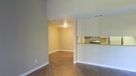 This single-story space showcases a practical and open interior layout designed for comfortable everyday living. The main living area features rich, dark wood-style flooring that extends throughout, providing a warm and cohesive foundation across the space. Neutral gray and beige tones on the walls create a calm, versatile backdrop suitable for a variety of furnishing styles.

A wide open passageway connects the main living area to an adjacent room, which shares the same flooring and warm beige wall tones, maintaining visual continuity throughout. This secondary space benefits from its own ceiling-mounted light fixture, ensuring adequate illumination.

To the right of the main living area, a kitchen is partially visible through a pass-through counter opening framed by a striped accent wall treatment. The kitchen features cabinetry and counter space, with a stainless steel appliance visible from the living area. The pass-through design fosters an open, connected feel between the kitchen and living spaces.

White baseboards trim the perimeter of each room, adding a clean, finished look. Ceiling-mounted HVAC vents are present throughout, ensuring climate control. Overall, the space presents a functional and well-connected layout with consistent finishes.