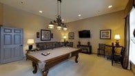 This single-floor space is designed as a dedicated entertainment and recreation area, centered around a full-sized pool table with ornate carved wood legs and a neutral felt surface. An elegant wrought-iron pendant light fixture hangs directly above the table, providing focused illumination while adding a decorative focal point to the room.

The walls are finished in a warm neutral tone, creating a welcoming and relaxed atmosphere. A wall-mounted flat-screen television is positioned prominently on one wall, making the space suitable for both gaming and viewing. A comfortable upholstered sofa with decorative pillows is arranged nearby, offering casual seating for spectators or guests.

Additional seating is provided throughout the room, including side chairs and a small accent table with a table lamp, contributing to a cozy ambiance. Decorative framed artwork is displayed on multiple walls, enhancing the refined yet casual character of the space. A low media console provides surface space along one wall, adding functional storage. Recessed ceiling lighting supplements the pendant fixture, ensuring the room is evenly and warmly lit throughout.