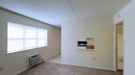 This single-floor space showcases a straightforward, functional layout with a neutral color palette throughout. The living area is finished with wall-to-wall carpeting and features a window fitted with horizontal blinds, allowing natural light to brighten the room. Beneath the window sits a wall-mounted HVAC unit, providing climate control directly within the living space.

A defining architectural detail is the pass-through window cut into the dividing wall, offering a direct view into the kitchen area. Through this opening, a gas range and wooden cabinetry are visible, suggesting a compact but practical kitchen layout. This pass-through design connects the two spaces without fully opening the floor plan, maintaining a sense of separation while promoting communication between areas.

The walls are finished in a clean, off-white tone, contributing to a bright and understated ambiance. Baseboard trim runs consistently throughout the space, adding a clean, finished look. The overall design is minimal and utilitarian, making it a practical space well-suited for efficient, everyday living.