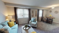 This single-floor residence offers a thoughtfully arranged layout that balances comfort and functionality. The open living area is generously sized, featuring soft carpeted flooring, neutral gray walls, and multiple windows dressed with layered blinds and drapes that invite an abundance of natural light. The space flows seamlessly into a dedicated dining area, where a round wood-top table and upholstered chairs create an intimate setting beneath a softly lit ceiling fixture.

The kitchen is well-equipped with warm wood cabinetry extending to the ceiling, offering ample storage. Granite-toned laminate countertops provide a practical workspace, complemented by a stainless steel sink and black appliances, including a gas range.

The bedroom is a serene retreat, carpeted and bathed in soft natural light through a window framed by tasteful drapes. It features a walk-in closet accessible through a dedicated doorway, offering generous storage. The adjoining full bathroom is finished with tile flooring, warm wood cabinetry, a white vanity with a large mirror, and bright overhead lighting, creating a clean and functional space.

Throughout the residence, a cohesive palette of soft grays, warm neutrals, and accent tones creates a calm and welcoming ambiance.