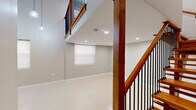 Located at 36 Third St, Unit 105, in Fall River, MA, this two-floor unit offers a thoughtfully designed layout spread across two distinct levels. The main floor impresses immediately with its soaring double-height ceilings, creating a dramatic sense of vertical space. Polished tile flooring reflects natural light admitted through multiple windows fitted with roller shades, while pendant lighting adds a modern touch overhead. A sleek recessed-lit lower area provides additional functional space beneath the upper level overhang. The staircase is a standout architectural feature — crafted from rich warm-toned wood with dark metal balusters and sturdy newel posts, it connects both floors with style and character. A wood-paneled entry door grounds the lower level with a welcoming, solid presence. The upper floor offers an open, light-filled space with smooth luxury vinyl flooring, recessed ceiling lights, and a large window that frames views of the surrounding neighborhood. A wood-and-iron railing lines the open mezzanine edge, maintaining visual connectivity with the floor below. The bathroom is finished to a high standard, featuring large-format tile flooring, a dark wood vanity with granite countertop, a backlit mirror, and a walk-in shower with white subway tile surround.