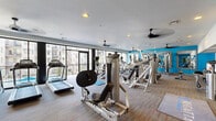 Located at 1565 Raleigh St in Denver, CO, this fitness center at the Regatta community is a well-appointed, single-floor facility designed to support a comprehensive workout experience. The space is thoughtfully laid out to accommodate a wide range of exercise needs, from cardiovascular training to strength conditioning.

The cardio section features a row of Matrix treadmills, elliptical trainers, and stationary bikes, all positioned along expansive floor-to-ceiling windows that flood the room with natural light and offer views of the surrounding community. Ceiling fans are distributed throughout the space, enhancing ventilation and comfort during workouts.

The strength training area is equipped with a variety of Life Fitness and Matrix selectorized machines, including leg press, cable systems, and multi-purpose weight stations. A dedicated free weights section includes a rack of dumbbells and a bench press with a barbell rack. Bold turquoise accent walls add a vibrant, energetic atmosphere to the space, complemented by large framed mirrors that visually expand the room and assist with exercise form.

The flooring consists of durable, multi-toned carpet tiles suited for a high-traffic fitness environment. The facility is accessed through double glass doors at the entrance, which are clearly marked. Recessed lighting throughout ensures consistent illumination across all workout zones.