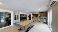 This single-floor recreational space at Viera at the Palm Beaches is thoughtfully designed for social gatherings and leisure. The centerpiece of the room is a full-size pool table with a tan felt surface and a sleek industrial-style base, positioned on light-toned tile flooring that runs throughout the space. A wall-mounted cue rack keeps the area organized and ready for play.

One of the most striking design elements is the reclaimed wood accent wall, composed of multi-colored planks in earthy tones of tan, grey, and blue, providing a bold visual contrast to the otherwise neutral palette of the room. Mounted on this wall is a flat-screen television, making the space equally suited for watching sports or entertainment.

Adjacent to the accent wall is a compact wet bar area, outfitted with light wood cabinetry, a stainless steel countertop, white subway tile backsplash, upper storage cabinets, and a glass-door beverage refrigerator — ideal for refreshments during gatherings.

The room is bathed in natural light, courtesy of multiple large sliding glass doors and windows that open to lush outdoor greenery and a paved patio area, creating a seamless indoor-outdoor connection. The beadboard ceiling adds a classic architectural touch, while recessed lighting ensures the space is well-lit at any hour.
