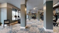 Located at 1775 Ashlar Dr in Fort Collins, CO, this well-appointed fitness center offers a generously sized exercise space designed to support a comprehensive workout experience. The layout is open and spacious, anchored by a series of structural columns that define distinct workout zones without enclosing the room, maintaining an airy, welcoming atmosphere throughout.

The cardio section is positioned along a wall of large, floor-to-ceiling windows that flood the space with natural light and provide views of the surrounding greenery. This area is equipped with treadmills, ellipticals, stationary bikes, and a stair climber. Ceiling fans overhead ensure comfortable airflow during workouts.

The strength training area features a broad selection of cable machines, weight benches, free weights, and resistance equipment. Wall-mounted televisions are placed throughout for entertainment. Full-length mirrors line portions of the walls, enhancing the sense of space and allowing users to monitor their form.

Underfoot, a bold blue-and-cream patterned carpet adds a stylish, modern touch to the interior. The walls are finished in a cool sage-gray palette, complemented by warm wood accent paneling in select areas. Exposed ductwork in portions of the space lends an industrial character. Recessed and overhead lighting ensures the entire floor is well-illuminated, creating a functional and inviting environment for all fitness levels.