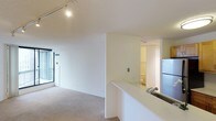 This single-floor space is designed with an open-concept layout that integrates the kitchen and living area into one cohesive environment. The living area features neutral-toned carpet flooring and white walls, creating a bright, clean atmosphere. A large window allows natural light to filter in, enhancing the sense of openness. Track lighting mounted on the ceiling provides additional illumination and a modern touch.

The kitchen is positioned adjacent to the living space, separated by a white countertop peninsula that serves as both a workspace and a visual divider. A stainless steel sink is built into the counter, and a refrigerator is visible nearby, indicating a functional kitchen setup. The white cabinetry and countertops maintain the space's consistent, minimalist aesthetic.

Visible from the main area, a bathroom is accessible through an open doorway, featuring tile flooring and a toilet, suggesting a practical and efficiently designed layout. The overall ambiance of the space is clean and understated, with white walls and light finishes throughout lending a bright, neutral backdrop suitable for a variety of uses.