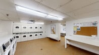 Located at 555 Sherbourne Street in Toronto, this shared laundry facility offers a practical and well-organized amenity space across a single floor. The facility is divided into two laundry rooms, both finished with white tile walls and durable small-format tile flooring that is easy to maintain. The first laundry room is generously sized and houses a large bank of stacked washer-dryer units along one wall, providing substantial capacity for residents. Additional front-loading washing machines line another wall, accompanied by long folding tables for sorting and preparing laundry. Informational bulletin boards are mounted on the walls for resident notices. The second laundry room features more stacked washer-dryer units and additional folding table space, along with a utility sink and further posted information boards. Both rooms are brightly lit with overhead fluorescent lighting and equipped with ventilation systems in the ceilings. Connecting the two laundry areas is a corridor that has been thoughtfully appointed as a waiting lounge, featuring a pair of comfortable upholstered armchairs, a small bookshelf stocked with reading materials, and a large, vibrant yellow and white canvas artwork adorning the wall — adding a welcoming touch to the functional space.