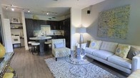 This thoughtfully designed single-floor space seamlessly blends comfort and modern style in an open-concept layout. The living area serves as the heart of the home, featuring a plush white sofa accented with blue and yellow throw pillows, a patterned accent chair, and a glass-top coffee table resting atop a textured area rug. A striking decorative candelabra centerpiece and coordinating wall art add artistic flair to the neutral gray walls, while warm ambient lighting from a floor lamp enhances the inviting atmosphere.

The kitchen is a standout feature, showcasing rich dark espresso cabinetry that extends to the ceiling, providing generous storage. A curved breakfast bar with bar stool seating offers a casual dining option, while the sleek countertops and tile backsplash add a polished finish. Track lighting overhead ensures the kitchen is well-lit and functional.

The warm-toned hardwood flooring flows continuously throughout the space, tying together the living and kitchen areas in a cohesive and elegant manner. The overall ambiance is contemporary and curated, with a neutral color palette punctuated by yellow and blue accents that give the space a fresh, modern energy.