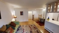 This single-floor space offers an open and airy layout that seamlessly connects its key living areas. Warm hardwood flooring flows continuously throughout, lending a cohesive and inviting feel to the entire space.

The living area is generously proportioned, featuring clean white walls that maximize the sense of brightness. A ceiling fan provides both comfort and ambient lighting overhead. Decorative touches, including a framed artwork piece on the wall and a boldly patterned area rug, add personality and warmth to the space.

Toward the rear of the space, a dining nook provides an intimate setting for meals, accompanied by a small table and chairs positioned beneath a ceiling light fixture. Nearby, a staircase with a classic wooden newel post and railing offers access to a lower level, suggesting additional usable space below.

The layout is practical and well-organized, with a hallway leading to what appears to be a closet or utility area with paneled doors. White baseboard trim throughout adds a clean, finished detail to the walls. The overall ambiance is comfortable and functional, with a neutral, welcoming aesthetic that provides a versatile foundation for a variety of interior styles.