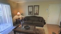 This single-floor space features a traditionally styled living room that exudes a warm and inviting ambiance. The room is finished with neutral, earthy-toned walls that create a calming backdrop for the classic furnishings throughout. A generously sized, dark upholstered sofa serves as the centerpiece of the seating area, accompanied by a wooden coffee table with a glass inset top, ideal for both function and display. A matching wooden end table sits beside the sofa, topped with a warm-toned table lamp that contributes to the room's soft, comfortable lighting. Decorative touches, including a glass vase and a small bowl, are arranged thoughtfully on the coffee table. Two framed botanical prints hang symmetrically above the sofa, adding a refined, classic character to the wall. A lush indoor plant in the corner brings a subtle natural element to the space. The floor is covered with neutral carpet, layered with a patterned area rug that anchors the seating arrangement. A wall-mounted thermostat is visible, indicating modern climate control. Overall, the living room presents a tidy, well-appointed environment with a cohesive traditional aesthetic.