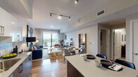 This thoughtfully designed single-floor residence showcases a seamless open-concept layout that connects the kitchen, dining, and living areas into one cohesive, light-filled space. Warm hardwood flooring flows throughout, complementing the clean, contemporary aesthetic. Track lighting in the main living area and recessed lighting elsewhere provide layered illumination across the space.

The kitchen is a standout feature, outfitted with rich walnut-toned lower cabinetry, crisp white upper cabinets, a classic subway tile backsplash, quartz countertops, and a full suite of stainless steel appliances. A kitchen island with seating extends into the dining area, creating a natural gathering point. The dining space transitions smoothly into the living room, which is anchored by a large sofa, curated artwork, and a set of sliding glass doors that open onto a private balcony overlooking a lush outdoor view.

The bedroom is a calm, well-proportioned retreat featuring soft neutral tones, bold artistic accents, and ample natural light from a large window. A dedicated media console and lounge chair add function and character to the room. The bathroom is clean and modern, featuring a wide vanity with an undermount sink, walnut cabinetry, a large mirror, and a shower-tub combination.