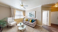This single-story residence offers a thoughtfully designed layout with a warm, neutral color palette and consistent crown molding detailing throughout. The living room serves as the central gathering space, featuring plush carpeting, a ceiling fan, and large windows dressed with layered curtains and blinds that invite generous natural light. A round white coffee table and a taupe sofa accented with teal and yellow throw pillows contribute to the room's inviting ambiance.

The bedroom quarters are tranquil and well-proportioned, each featuring ceiling fans, crown molding, and windows with sheer curtains that soften incoming light. One bedroom showcases a rich wood dresser, a decorative round mirror, and tasteful wall art, creating an elegant retreat.

The galley-style kitchen is efficiently organized with light wood cabinetry, white appliances including a refrigerator, electric range, and built-in microwave, all set atop wood-look vinyl flooring. A door at the end of the kitchen provides additional access.

The primary bathroom features a dual-sink vanity with light wood cabinetry and wood-look flooring, complemented by a walk-in closet. A second bathroom provides a single-sink vanity and a glass-enclosed shower stall, both finished with consistent, clean materials throughout.