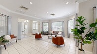 Located at Bristol Lakes in Mount Dora, Florida, this single-floor community clubhouse offers a bright and welcoming atmosphere designed for both relaxation and social gatherings. The space is finished with large white ceramic tile flooring throughout, crown molding, and recessed lighting that complement the neutral warm-gray walls.

The entrance welcomes guests with an elegant foyer featuring a mirrored console table, decorative artwork, and a cushioned accent bench beside a sun-filled window. Classic white French doors with glass pane detailing lead to and from the outdoor area, offering views of the surrounding grounds.

The open main lounge area is generously sized, featuring multiple seating arrangements with bold cognac leather chairs, a gray sofa with accent pillows, and a central coffee table, all anchored by a textured area rug. Large windows fitted with blinds and sheer curtains flood the space with abundant natural light.

A dedicated dining area accommodates a large round dark-top table surrounded by mixed wood-and-metal chairs, ideal for community meetings or events. Adjacent to it, a functional kitchen includes dark espresso cabinetry, stainless steel appliances, a bright countertop bar with seating for multiple guests, and windows overlooking the outdoor space — completing this versatile, community-focused facility.