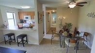 This single-story residence is designed with an open-concept floor plan that seamlessly connects the kitchen, dining, and living areas. The kitchen features warm wood-toned cabinetry, white appliances including a microwave, stove, and refrigerator, and a breakfast bar with bar stool seating — ideal for casual dining or entertaining. A pass-through counter connects the kitchen to the adjacent spaces, enhancing the open feel. The dining area accommodates a glass-top table with stylish metal chairs, complemented by a decorative indoor plant and accent pieces that add warmth to the room. A ceiling fan with a light fixture overhead provides both ventilation and ambient lighting. Neutral-toned carpeting flows throughout the main living areas, contributing to a soft, cohesive aesthetic. A hallway visible from the main living space leads to additional rooms, including a bedroom with light blue accent walls and natural light. The overall color palette of warm beige tones and wood accents creates a welcoming and functional atmosphere throughout the residence.