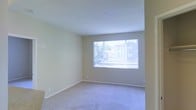 This single-floor space features a clean, open living area finished with wall-to-wall carpeting in a neutral tone that complements the light beige walls throughout. The room is characterized by its simplicity and functionality, with smooth white ceilings and clean baseboards framing the space neatly. A large picture window serves as a focal point on one wall, fitted with vertical blinds that allow for adjustable light control. The window draws in natural light, brightening the interior and providing a view of neighboring structures and surrounding greenery. The overall layout is open and uncluttered, offering a versatile blank canvas well-suited for a variety of furniture arrangements. The neutral color palette of the walls and flooring creates a cohesive, understated aesthetic throughout. A light switch and electrical outlets are positioned along the walls, and a recessed ceiling fixture provides additional overhead lighting. The space presents a straightforward, move-in-ready environment with a calm, functional ambiance.