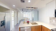 Located at 805 North Olive Avenue in West Palm Beach, Florida, this thoughtfully designed two-bedroom, two-bathroom residence offers a clean, contemporary aesthetic throughout its single-floor layout. The open-concept living and kitchen area serves as the heart of the home, where striking black track lighting contrasts beautifully against crisp white ceilings. The kitchen features a central island with quartz countertops, warm wood cabinetry, a stainless steel sink, and a stacked washer and dryer discreetly tucked behind louvered doors. Light wood-tone flooring flows seamlessly through the living spaces, enhancing the bright, airy atmosphere. The primary bedroom offers a generous footprint with natural light streaming through a large window that frames lush tropical palm views, complemented by a stylish barn door leading to a dedicated closet. The second bedroom is equally bright and well-proportioned. Both bathrooms are finished to a high standard, featuring frameless glass-enclosed walk-in showers, tiled surrounds, warm wood vanities, and white quartz surfaces. A connecting hallway and two closets round out the efficient floor plan, making this residence a polished and functional home in the heart of West Palm Beach.