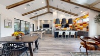 Located in Phoenix, AZ, this single-story community clubhouse offers a generously sized, open-plan gathering space designed for both socializing and recreation. The architecture is immediately striking, defined by soaring vaulted ceilings accented with exposed wood beams that lend a warm, rustic-modern character to the bright, airy interior. Large floor-to-ceiling glass panels and sliding doors flood the space with abundant natural light and provide seamless views of the surrounding outdoor areas.

The flooring throughout features wood-look tile in a neutral tone, providing a durable yet stylish foundation that unifies the various zones of the space. A central kitchen island with a crisp white quartz countertop and navy cabinetry serves as an eye-catching focal point, illuminated by three dramatic oversized pendant lights with gold interiors. Behind it, a vivid mosaic-style accent wall in bold shades of red, orange, and yellow energizes the room with color. Gray cabinetry, stainless steel appliances, and a mounted TV complete the fully equipped kitchen area.

The lounge zone features comfortable seating arrangements with a sofa, coffee table, and accent chairs, while a billiards table and multiple dining tables provide ample entertainment options. Potted olive trees add a natural, organic touch throughout, enhancing the welcoming ambiance of this well-appointed community amenity space.