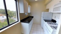 This single-floor space showcases a thoughtfully designed kitchen that balances functionality with clean, modern aesthetics. The kitchen is finished with striking dark granite countertops that contrast elegantly against the crisp white cabinetry, which offers generous storage through both upper and lower cabinets. White tile flooring runs throughout, contributing to a bright, cohesive look that enhances the sense of space. The kitchen is equipped with essential appliances, including a full-size white refrigerator and a stove, providing everything needed for everyday cooking. A stainless steel sink is positioned beneath a window, allowing natural light to filter in while offering a pleasant outdoor view — a subtle but welcome touch. Large windows along one wall further amplify the natural light, creating an open and airy atmosphere. The warm yellow-toned walls add a soft, inviting warmth to the space, complementing the clean white surfaces throughout. The layout is efficient and galley-style, maximizing every inch of available space while maintaining an uncluttered, practical flow. Overall, the kitchen presents a well-maintained and functional environment with a tidy, neutral aesthetic that is both welcoming and versatile.