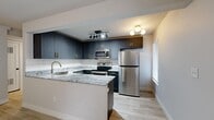 This efficiently designed two-bedroom, one-bathroom unit makes excellent use of its single-floor layout. The kitchen serves as a standout feature, showcasing slate-blue shaker cabinets with brushed gold hardware, granite countertops with striking veining, a white subway tile backsplash, and a full suite of stainless steel appliances including a refrigerator, electric range, and built-in microwave. A peninsula with a granite waterfall edge provides additional workspace and an open connection to the living area. The living room benefits from light wood-look luxury vinyl flooring and a set of French doors that introduce natural light and outdoor access.

The primary bedroom features the same wood-look flooring, a ceiling fan with a gold-toned fixture, and a wall-mounted mini-split HVAC unit for independent climate control. The second bedroom offers plush carpeting, its own ceiling fan, a window for natural light, and a wall-mounted mini-split unit. The bathroom is well-appointed with marble-look tile flooring, a subway-tiled shower/tub combo, and a navy vanity with granite countertop and an illuminated mirror. A stacked washer and dryer unit is conveniently integrated near the bathroom, maximizing the use of available space throughout.